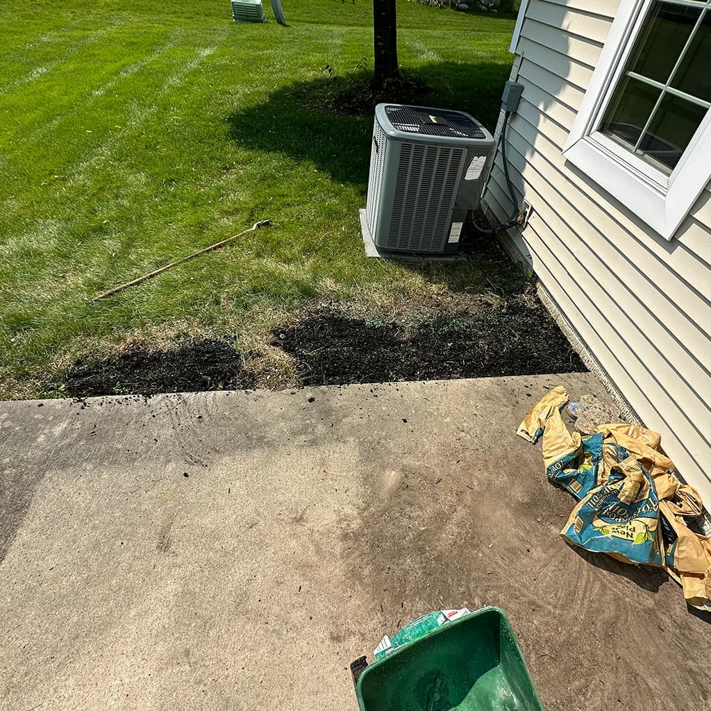 Lawn repair and overseeding prep at a Des Moines-area home: topsoil and seeding materials along a concrete walk beside the house, with a spreader and established lawn in view.